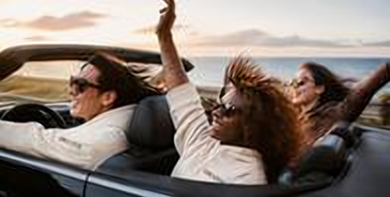 Friends in a convertible on a coastal highway at sunset, one passenger in a GROOVE hoodie